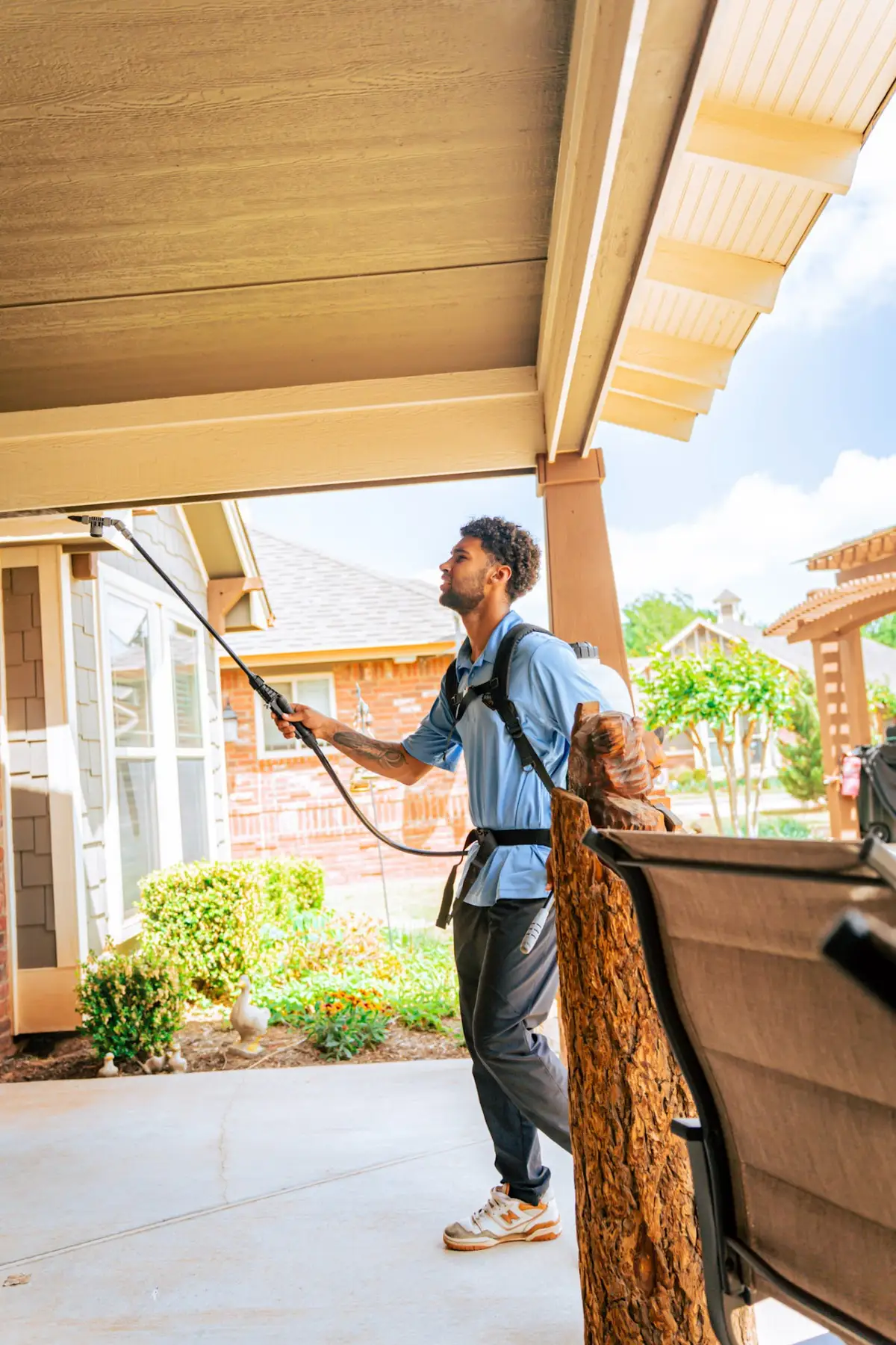 A technician wearing a backpack sprayer holds a long wand beneath the eaves of a house on a covered patio.