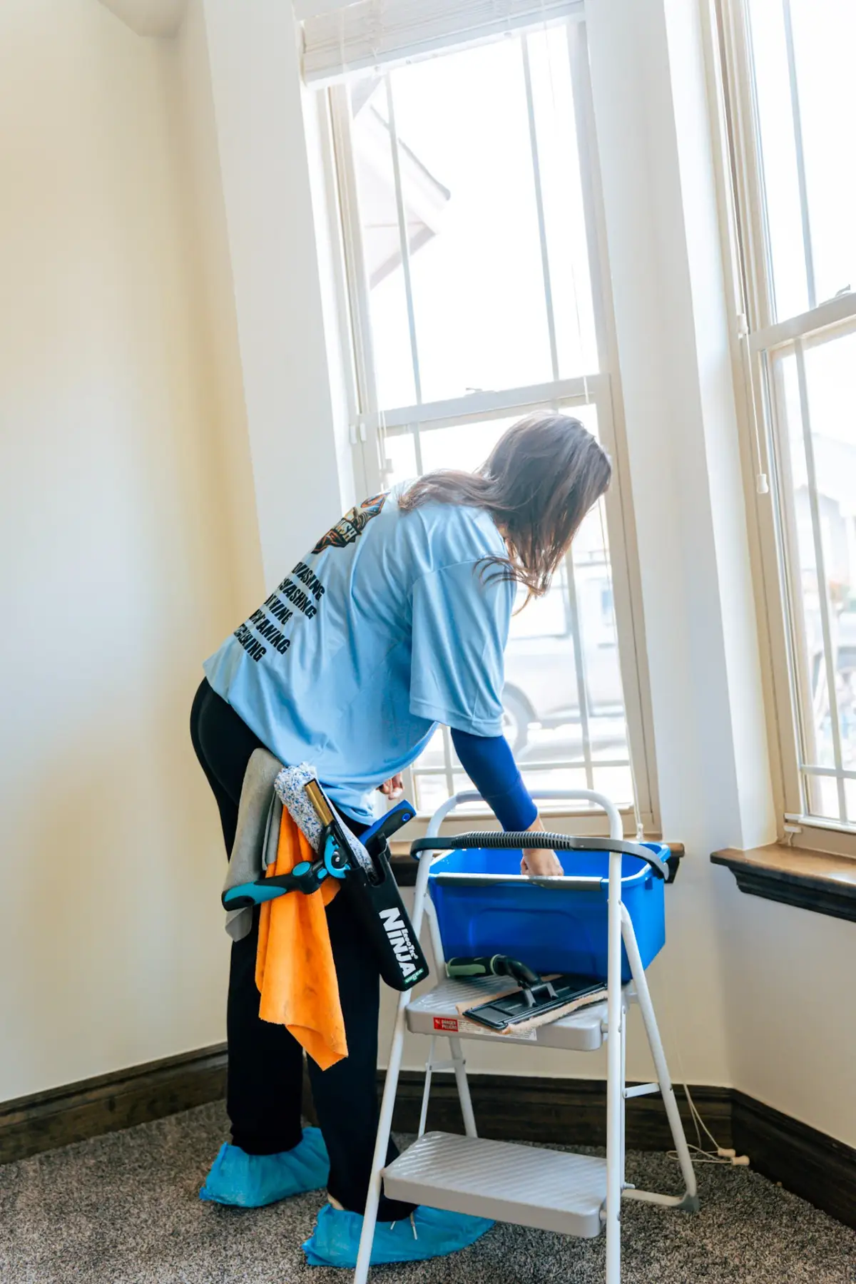 A worker in a light blue printed shirt and shoe covers reaches into a blue bucket on a stepladder beside indoor windows, with cleaning tools and towels hanging at their waist.