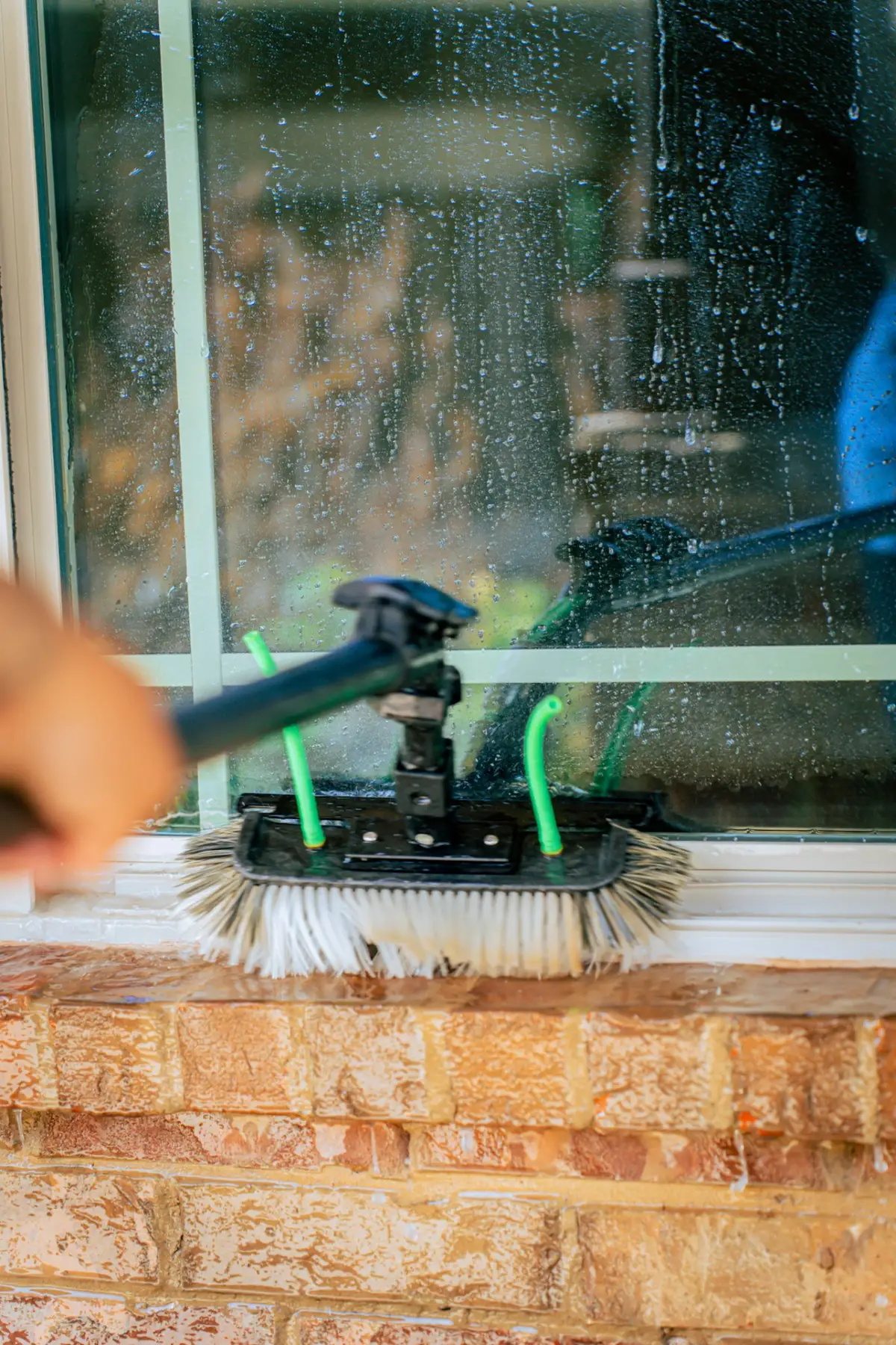 A pole-mounted brush with green water tubes cleans a wet exterior window above a brick sill. A person's hand holds the pole at the left edge.