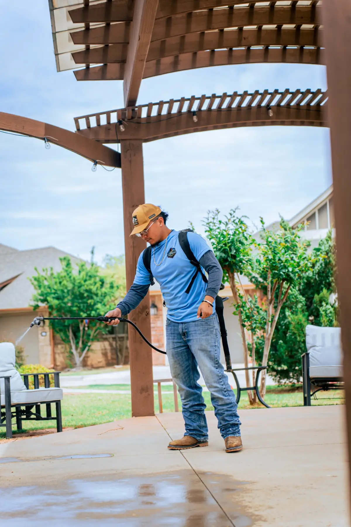 A worker wearing a backpack sprayer directs a spray wand beside patio seating beneath a wooden pergola.
