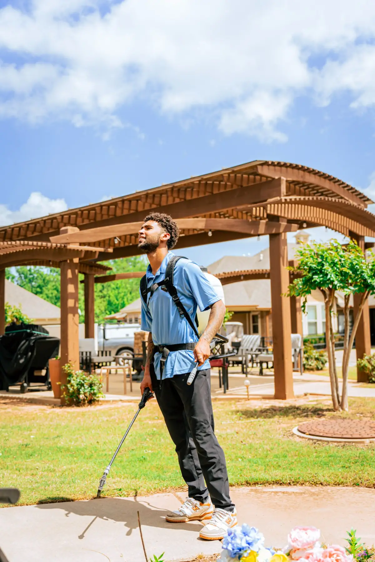 A worker in a blue polo carries a backpack sprayer and holds a long spray wand beside a lawn and patio with a wooden pergola.
