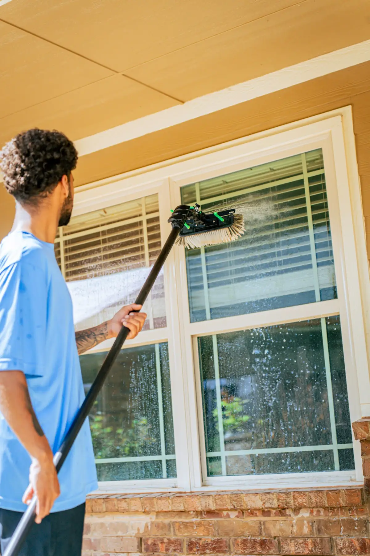 A worker in a blue shirt cleans exterior windows with a long-handled brush, with water spraying across the glass.