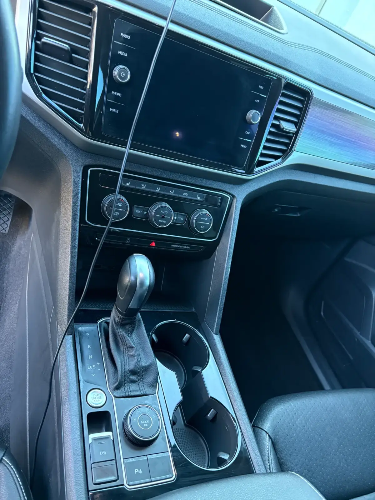Car interior showing a dark infotainment screen, dashboard vents, climate controls, automatic gear selector and two cup holders. A cable hangs across the dashboard and center console.