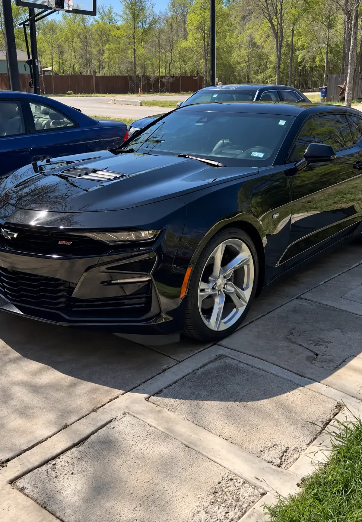 A glossy black Chevrolet Camaro parked on a concrete driveway, with other cars, a basketball hoop, and trees in the background.