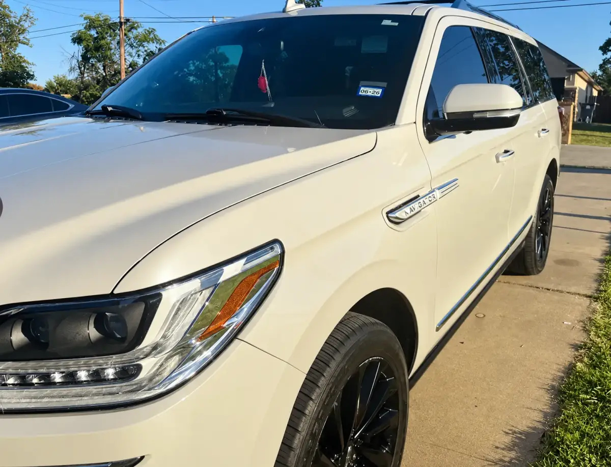 Close-up angled view of a white Lincoln SUV parked on a sunny driveway, showing its headlight, dark windows, side mirror and black wheels. No people or service-company branding are visible.