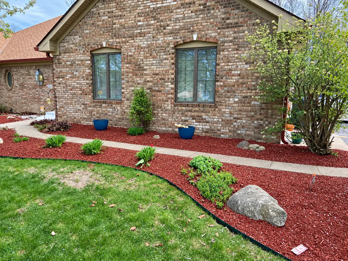 Red mulch beds with green plants, shrubs and decorative rocks border a concrete walkway outside a brick house. Blue planters sit beneath the windows, and a lawn fills the foreground.