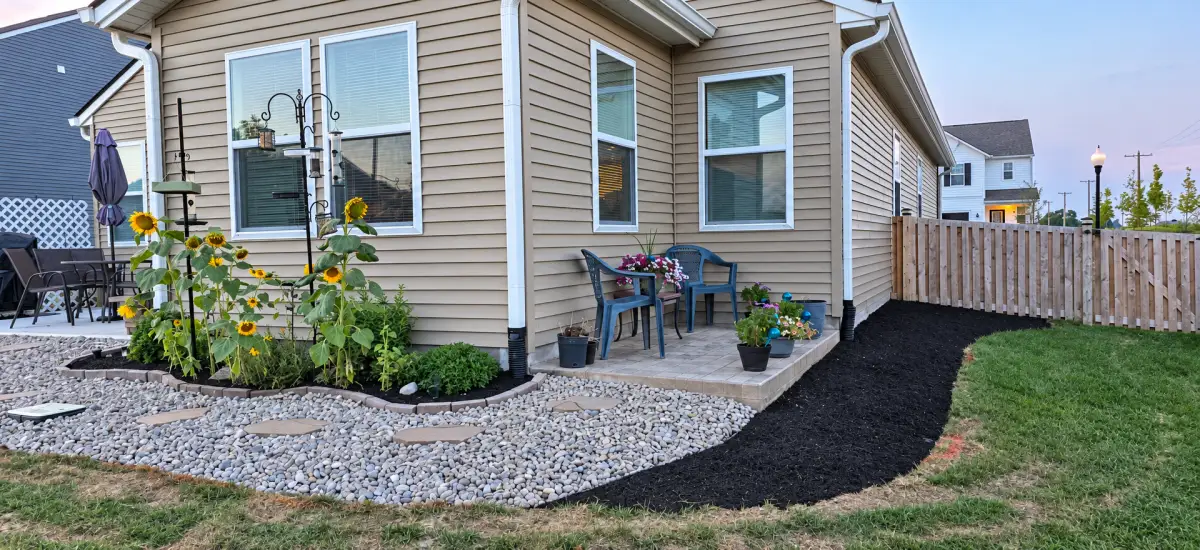 Backyard landscaping beside a house, with a curved black mulch bed, decorative stones and stepping stones, sunflowers, and a small patio with chairs and potted flowers.