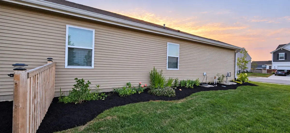 A curved garden bed with dark mulch, shrubs, and flowering plants runs along a beige-sided house beside a green lawn and wooden fence.
