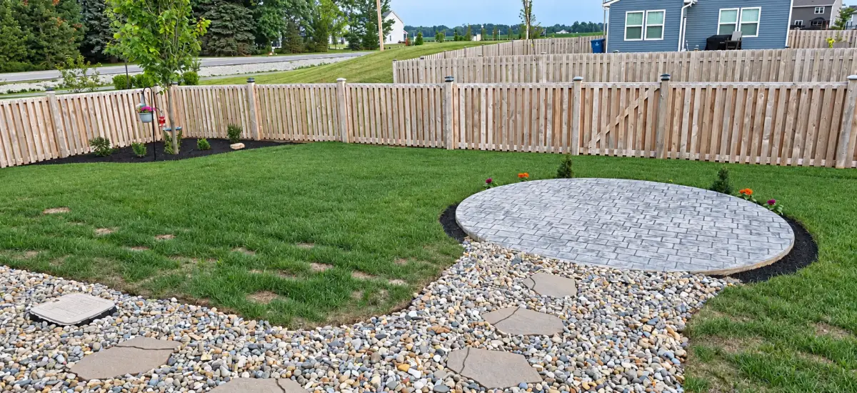 A circular gray stamped-concrete patio in a fenced backyard, with a river-rock and stepping-stone path, green lawn, and small planted beds.