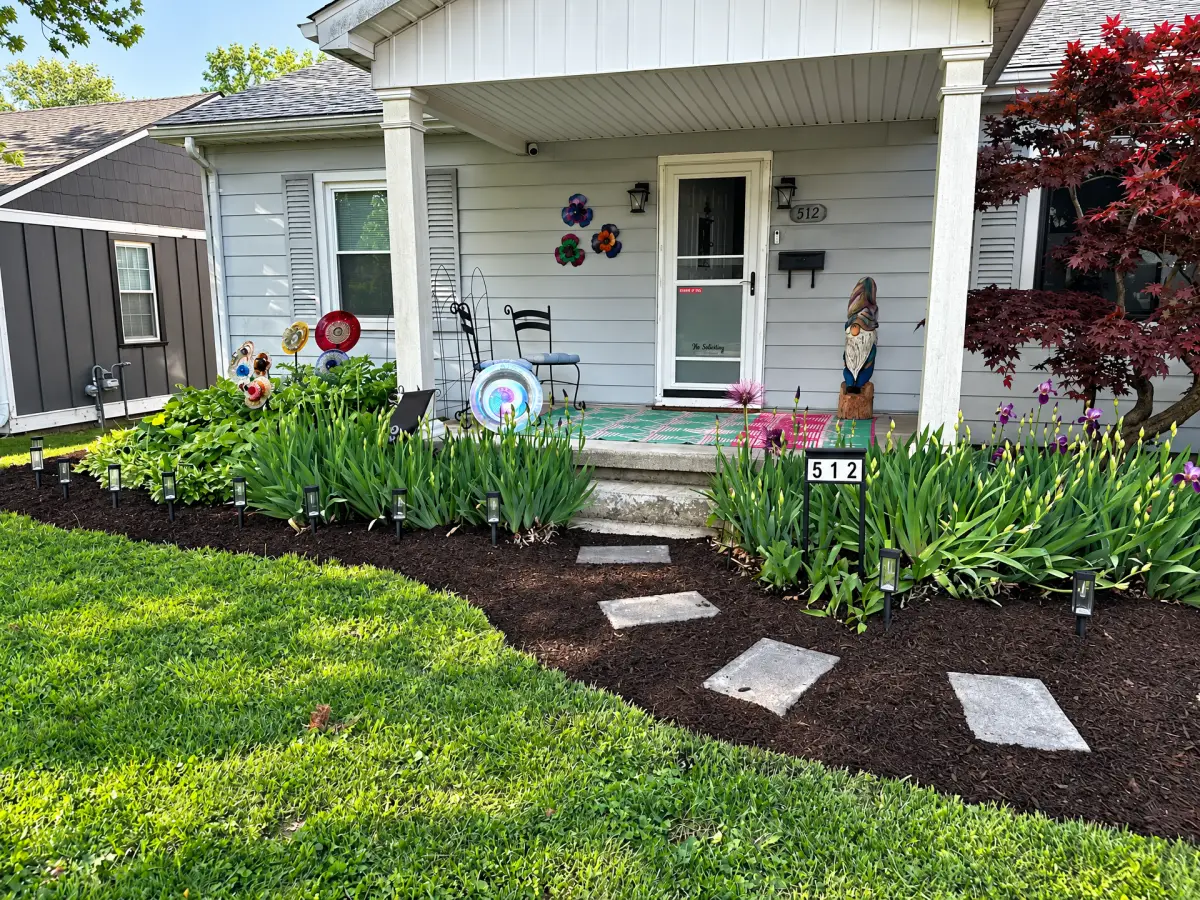 Front yard with dark mulch beds, flowering plants, small landscape lights, and stepping stones leading to a covered house porch.