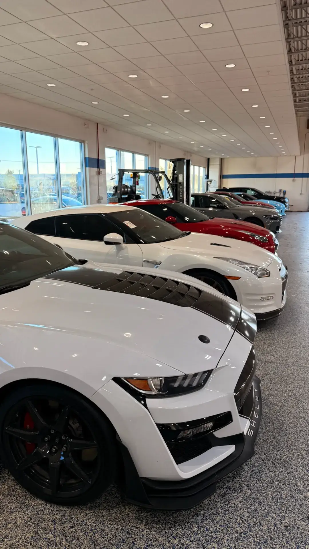 Sports cars parked in a row inside a bright automotive space with large windows, a speckled floor and a forklift behind the cars. A white car with black hood accents fills the foreground.
