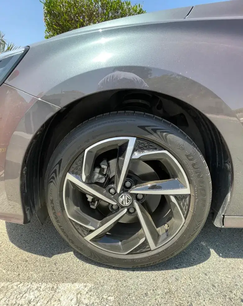 Close-up of a gray MG car’s front wheel, showing a Dunlop tire, alloy rim, brake components and surrounding fender outdoors.