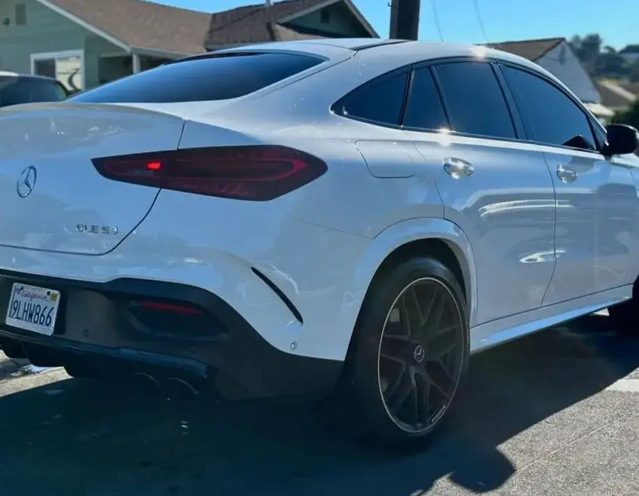 Rear and passenger-side view of a white Mercedes-Benz GLE 53 with dark tinted windows and black wheels, parked on a sunny residential street.