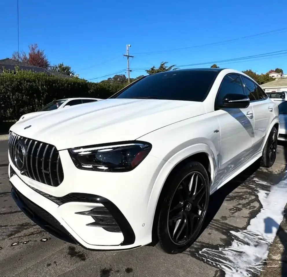 A white Mercedes-Benz SUV with black wheels and dark windows sits outdoors on wet pavement with soap suds beside it.