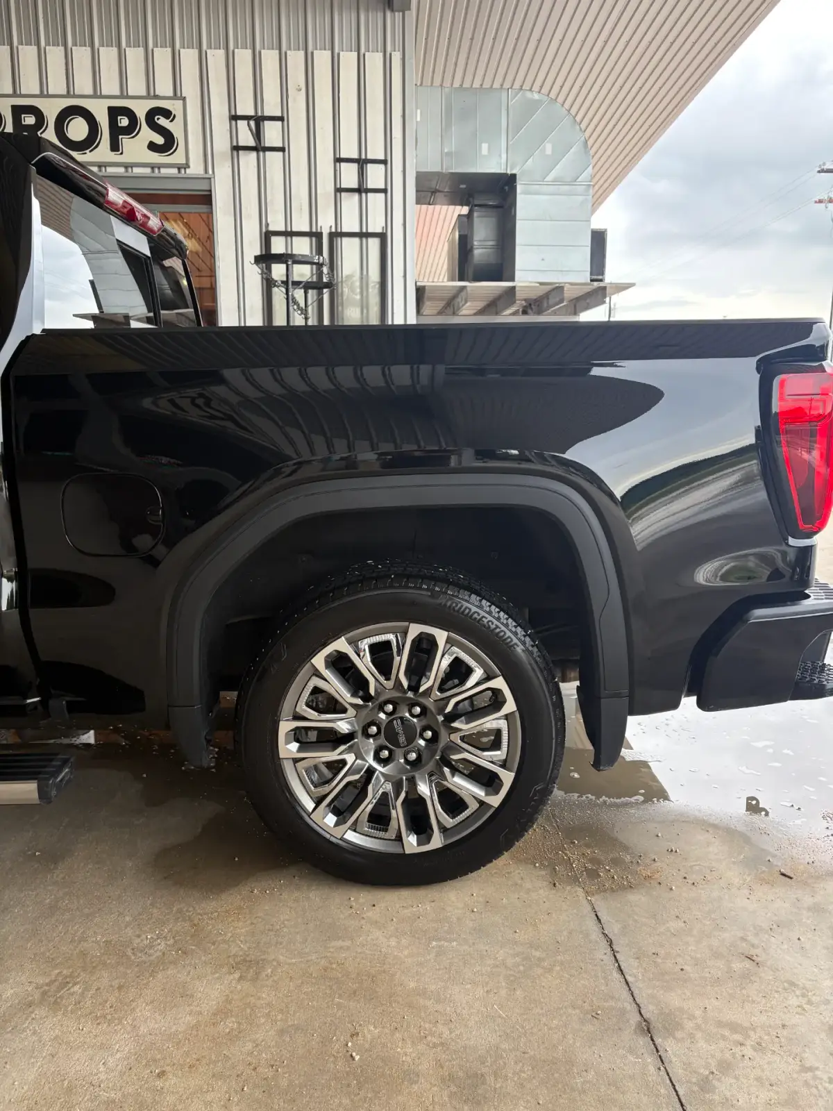 Rear side of a glossy black GMC pickup, showing its rear wheel, truck bed and red taillight beside a metal-sided building. Water and suds are visible on the concrete.