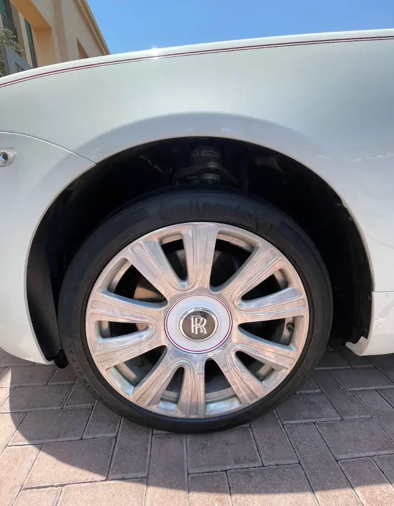 Close-up of a white Rolls-Royce’s wheel and fender, showing a silver multi-spoke rim, RR center emblem, and black tire on brick paving.