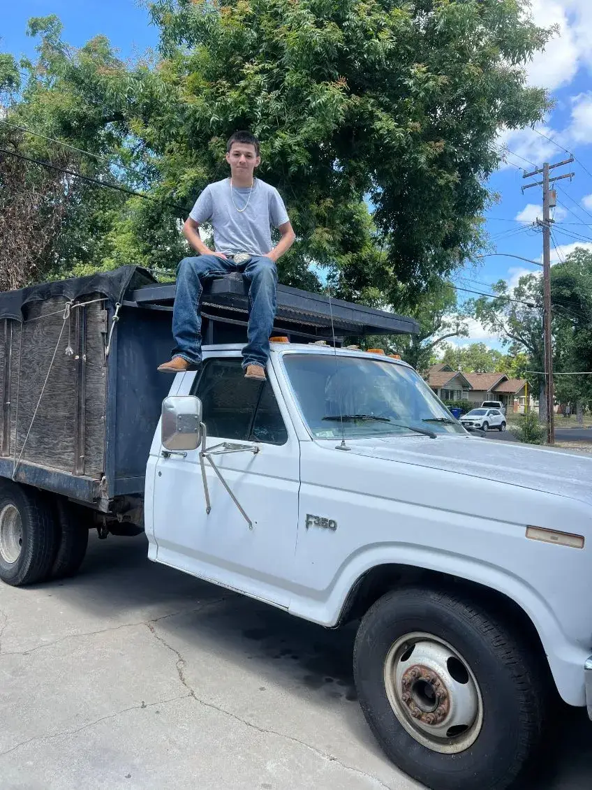 A person in a gray T-shirt and jeans sits above the cab of a white truck with tall dark cargo sides, parked outdoors near trees and houses.