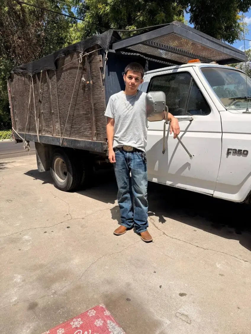 A person in a gray T-shirt and jeans stands beside a white Ford F350 truck with tall wooden cargo sides, a black tarp, and an overhead metal rack.
