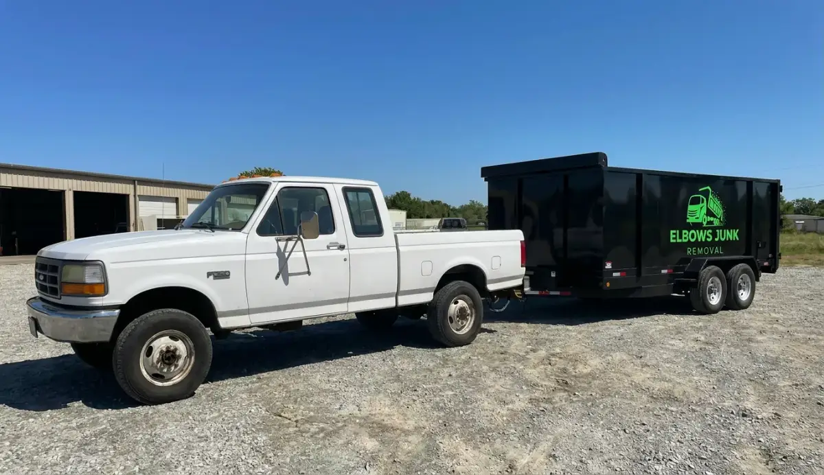 A white pickup truck attached to a black dump trailer with a green “ELBOWS JUNK REMOVAL” logo sits on a gravel lot outside garage buildings.