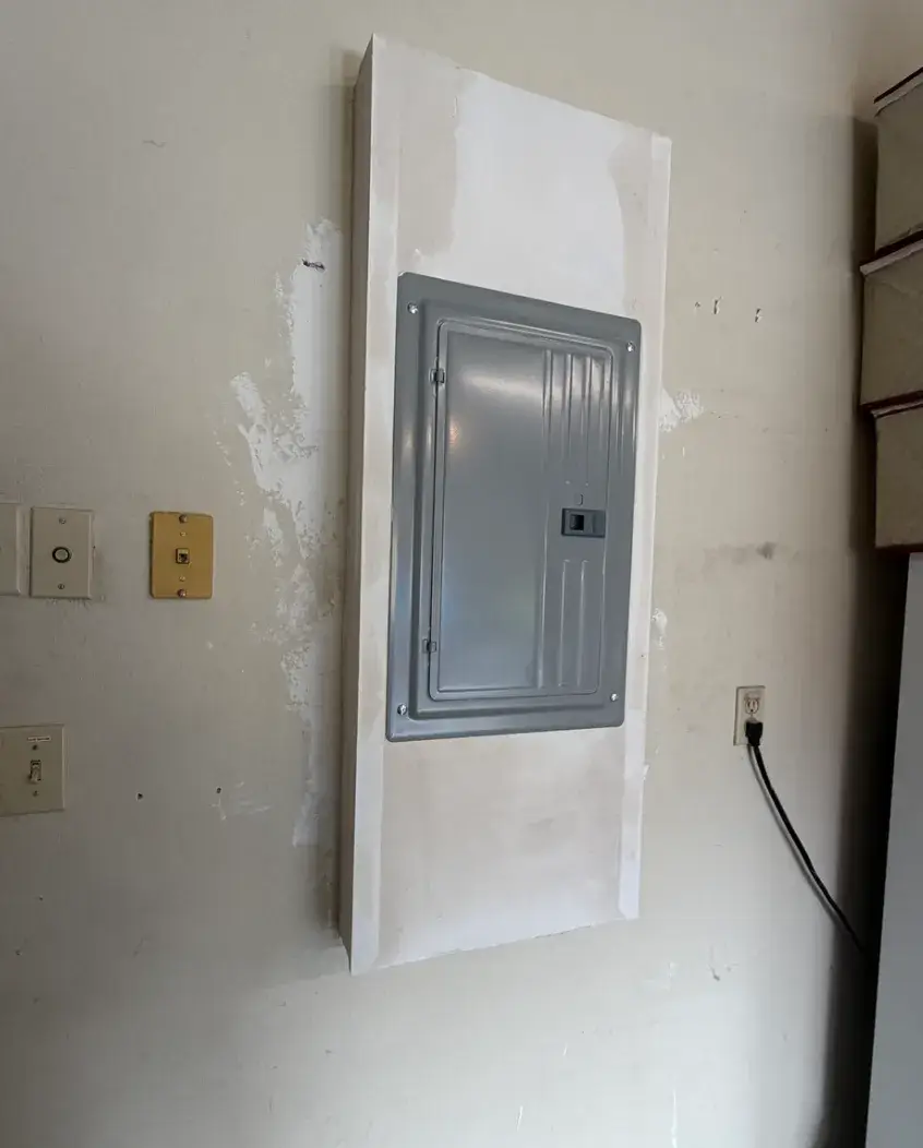A closed gray electrical panel mounted in an interior wall with unfinished white patching around it. Nearby wall plates and a plugged-in outlet are visible.