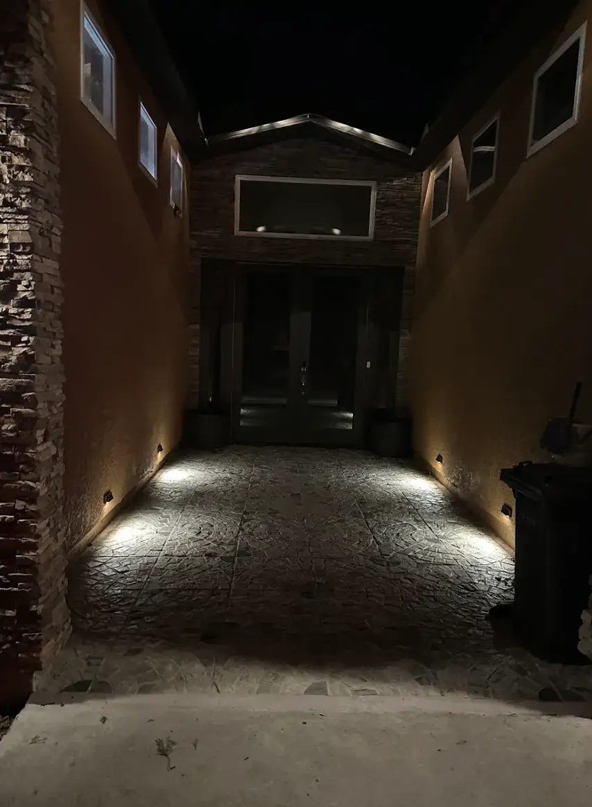 Nighttime view of a home entrance with low wall lights illuminating a textured walkway, stucco walls, stone accents, and dark double doors.
