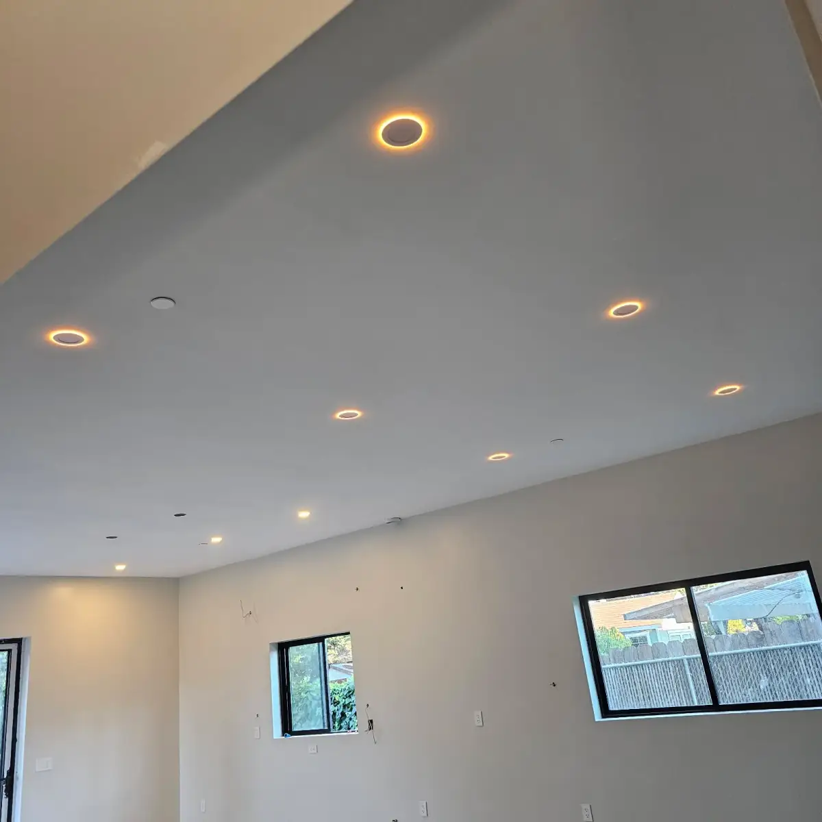 An unfinished room with illuminated recessed ceiling lights, black-framed windows, wall outlets, and exposed wires at several wall openings.