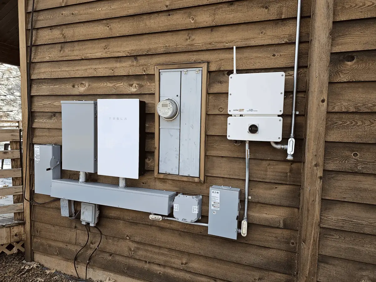 Tesla-branded electrical equipment, a SolarEdge inverter, a utility meter, and disconnect boxes mounted on a wood-sided exterior wall.