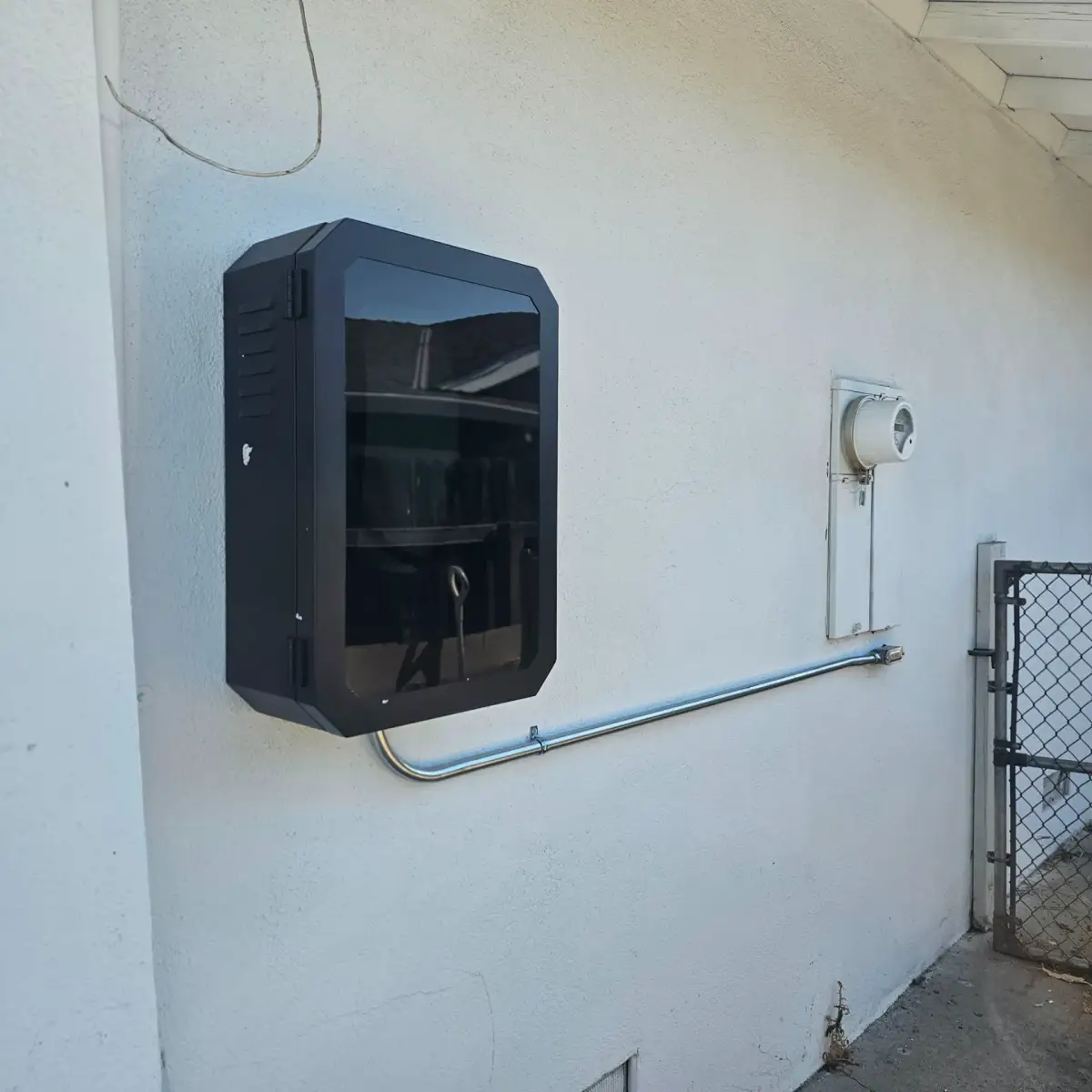 A black electrical enclosure mounted on a white exterior wall, with metal conduit running beneath it toward a nearby utility meter. A chain-link gate stands at the right.