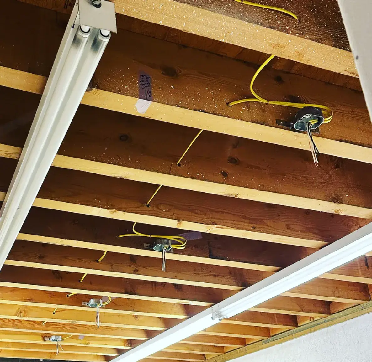 Exposed wooden ceiling joists with yellow electrical cables, open electrical boxes, hanging wire ends, and long fluorescent light fixtures.