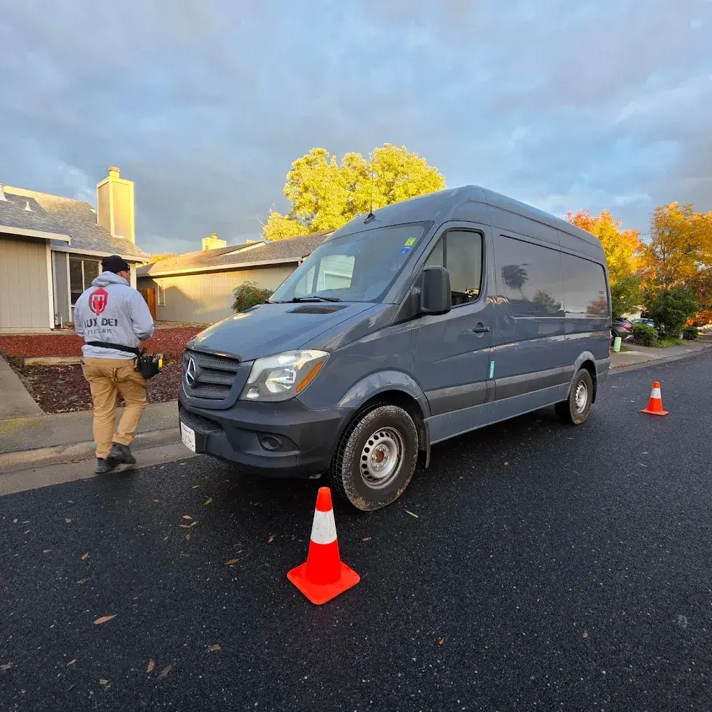 A worker wearing a logo hoodie and tool belt stands beside a gray cargo van on a residential street, with orange traffic cones nearby.