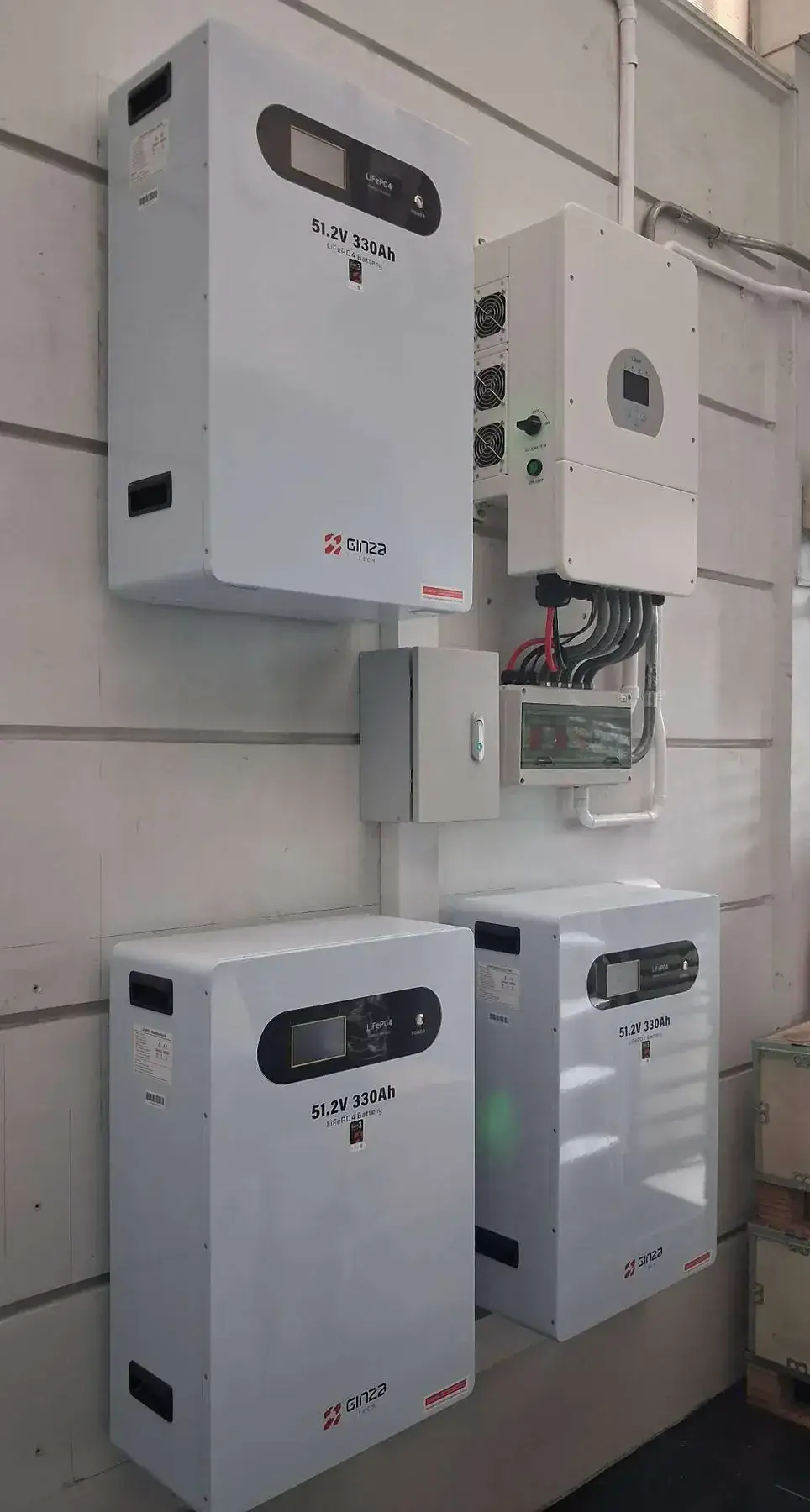 Three white lithium battery units mounted indoors beside an inverter, electrical boxes, and connecting cables. The batteries are labeled 51.2V 330Ah.