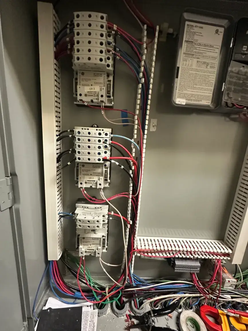 Inside an open electrical control cabinet, stacked contactors connect to red, black, blue and white wiring. Wire channels, terminal blocks, electrical tape and a partially visible clamp meter surround the components.