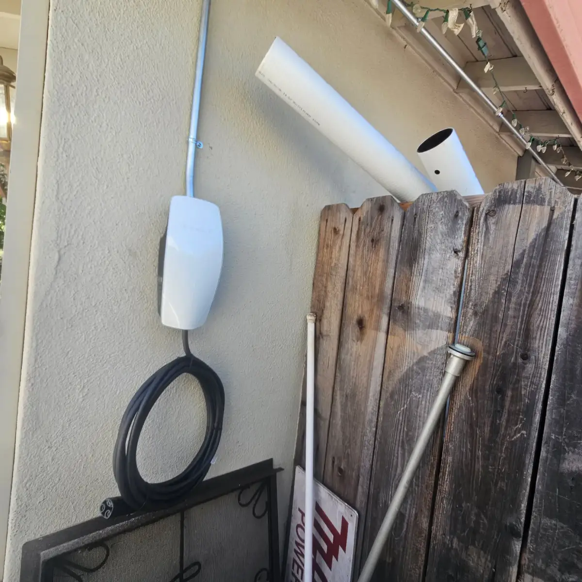A white Tesla electric vehicle charger is mounted on an exterior stucco wall, with metal conduit above and a coiled black charging cable below. A wooden fence and stored pipes stand beside it.