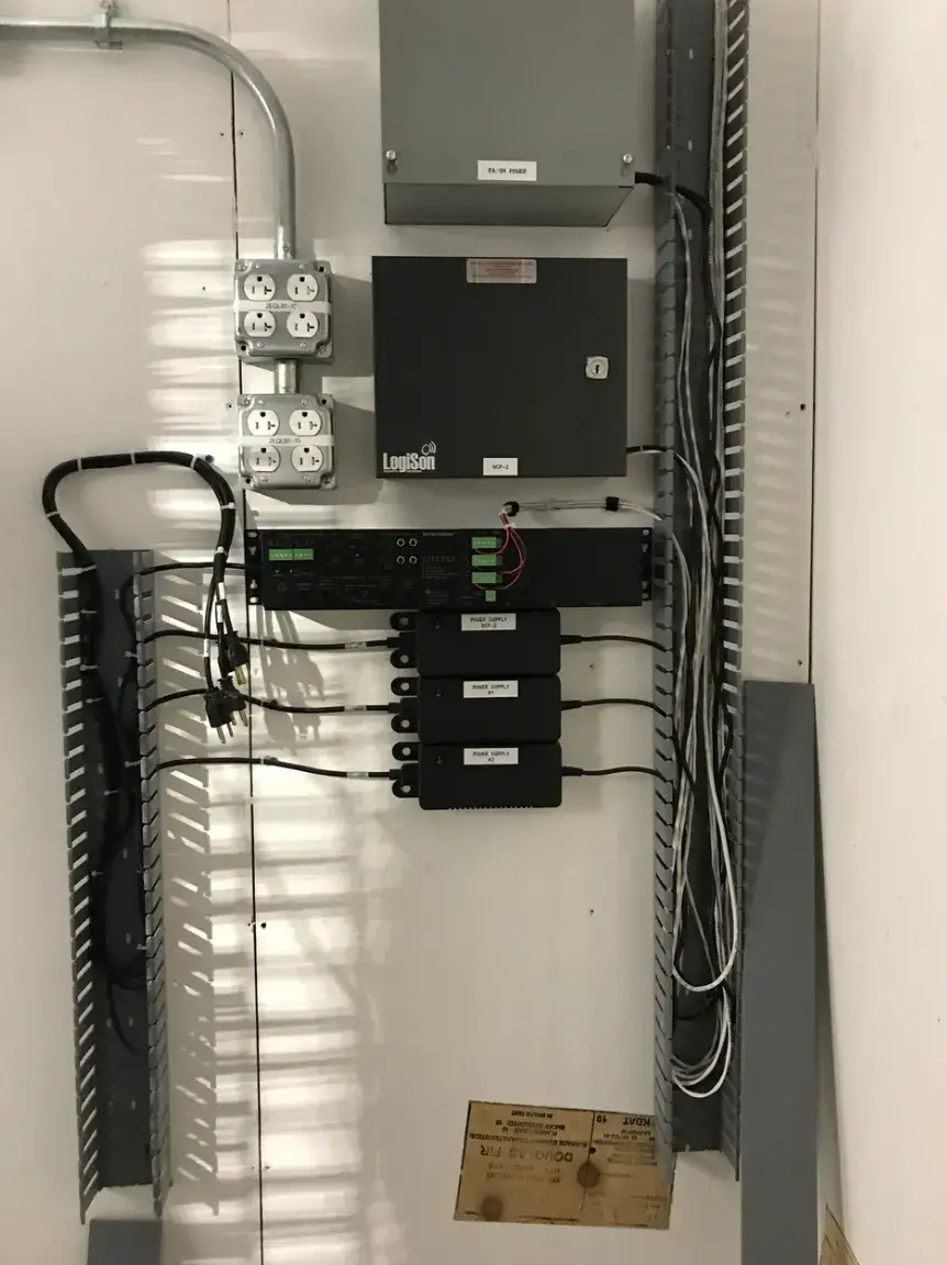 Wall-mounted electrical and audio control equipment, including a black LogiSon enclosure, outlets, labeled power supplies, conduit, and cables in open cable channels. No people are visible.