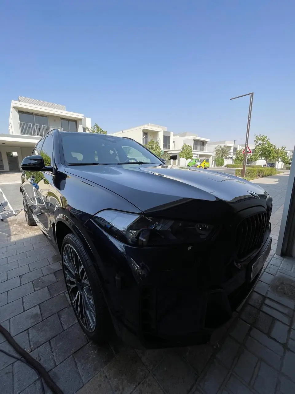 A glossy black BMW SUV parked on a paved residential driveway, viewed from the front corner, with modern homes behind it.
