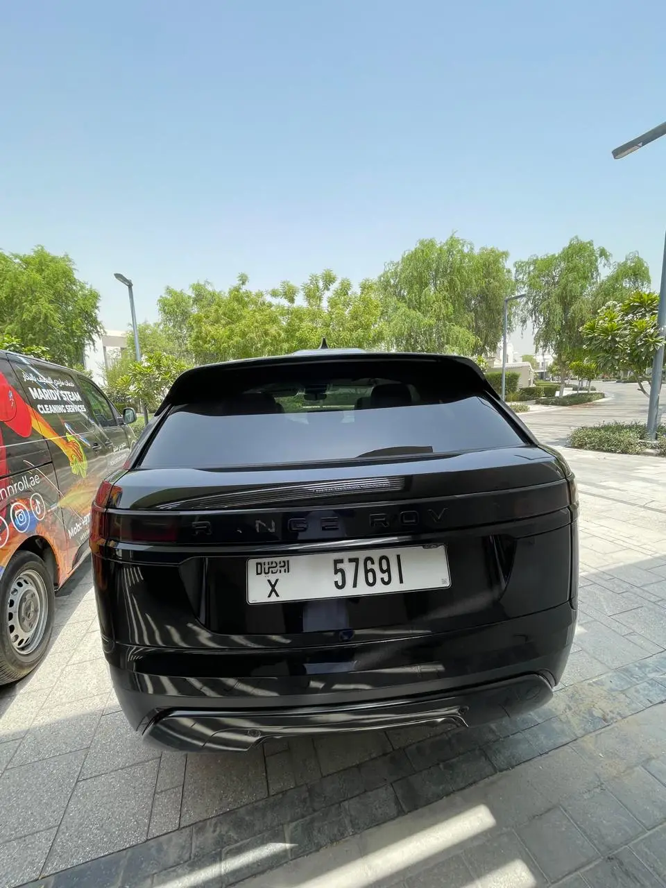 Rear view of a black Range Rover parked on a paved driveway beside a partially visible van branded for cleaning services, with trees in the background.