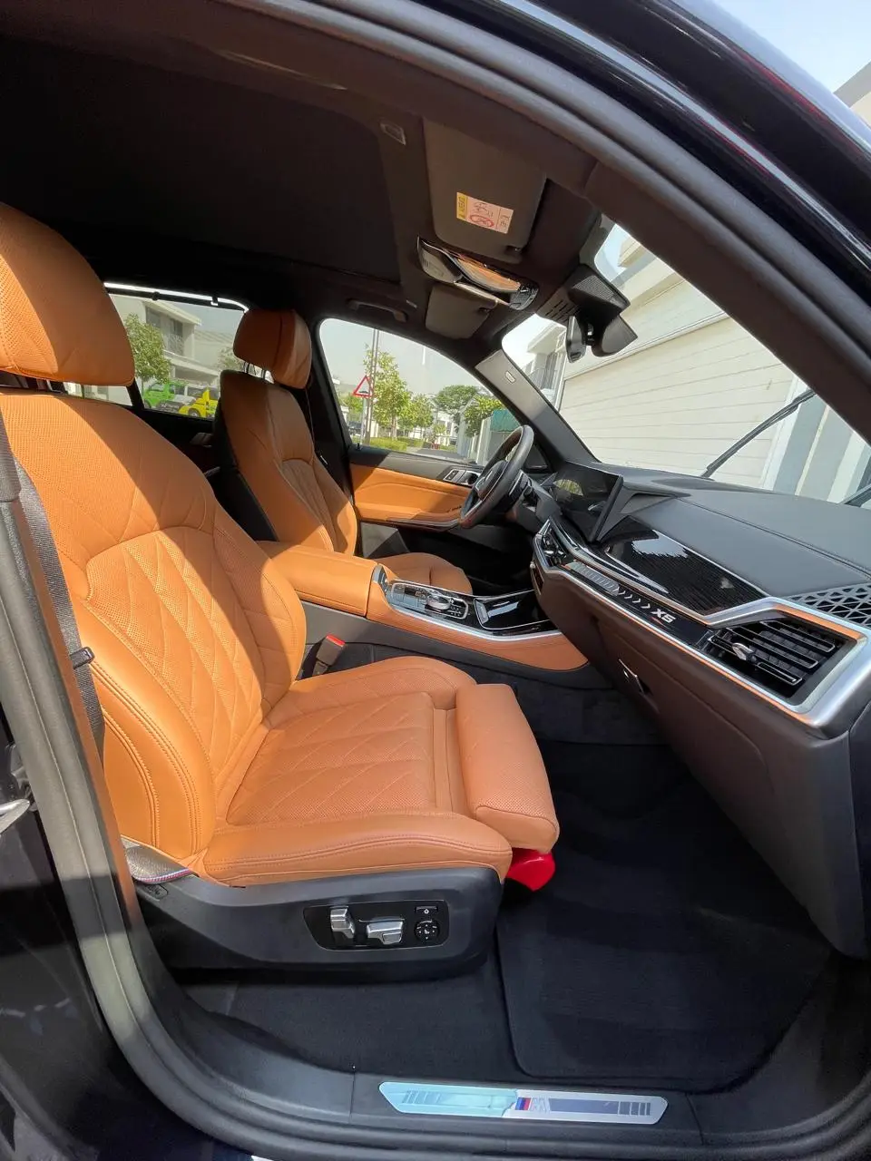View through an open passenger door of a BMW X5, showing tan quilted leather front seats, a black dashboard, center console, and dark floor mats.