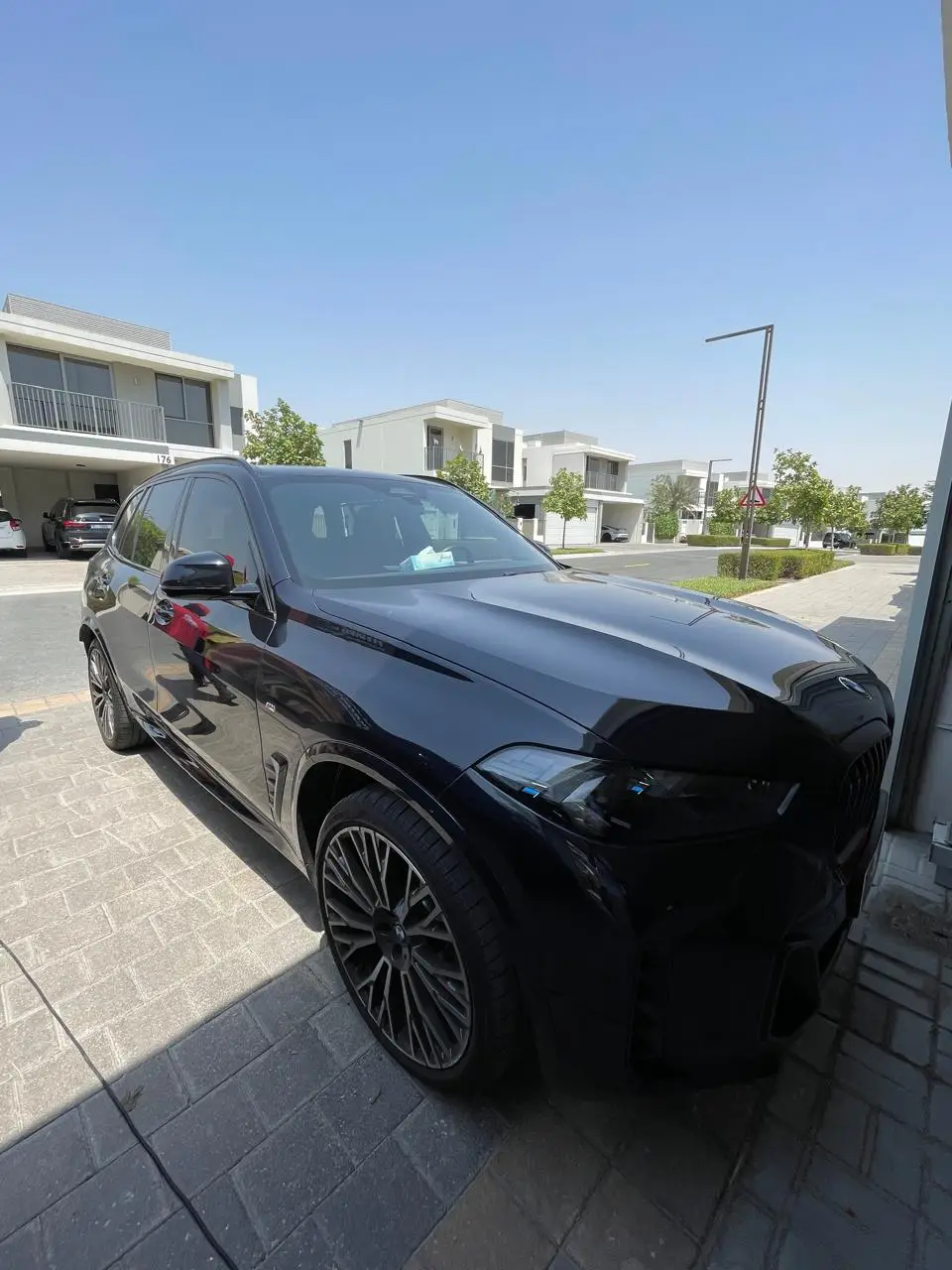 A glossy black BMW SUV parked on a paved driveway, viewed from the front corner, with modern homes across the street.