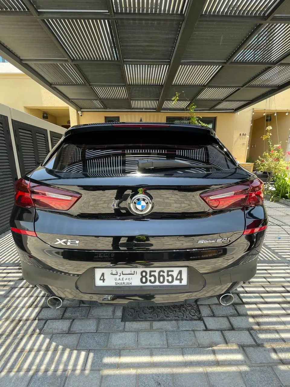 Rear view of a shiny black BMW X2 parked on a paved driveway beneath a slatted metal carport beside a yellow house.