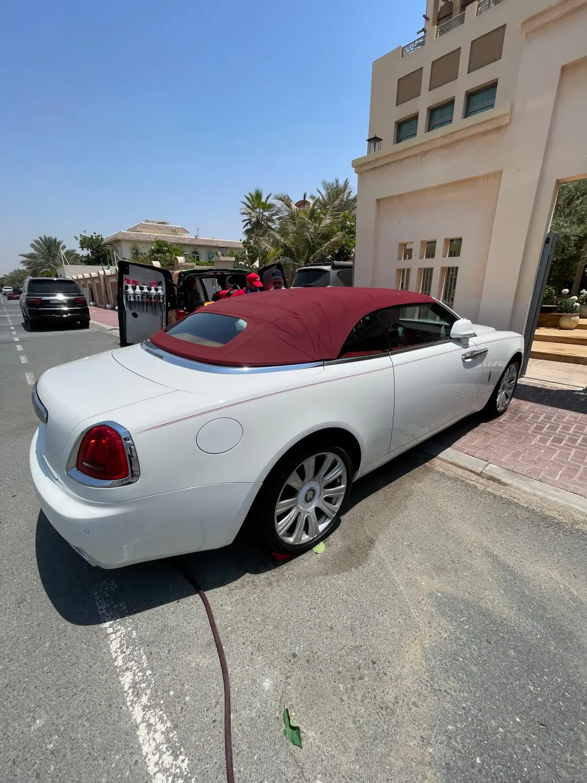 A white Rolls-Royce convertible with a burgundy soft top parked outside a residence. People stand beside an open vehicle stocked with spray bottles, and a hose runs across the pavement.